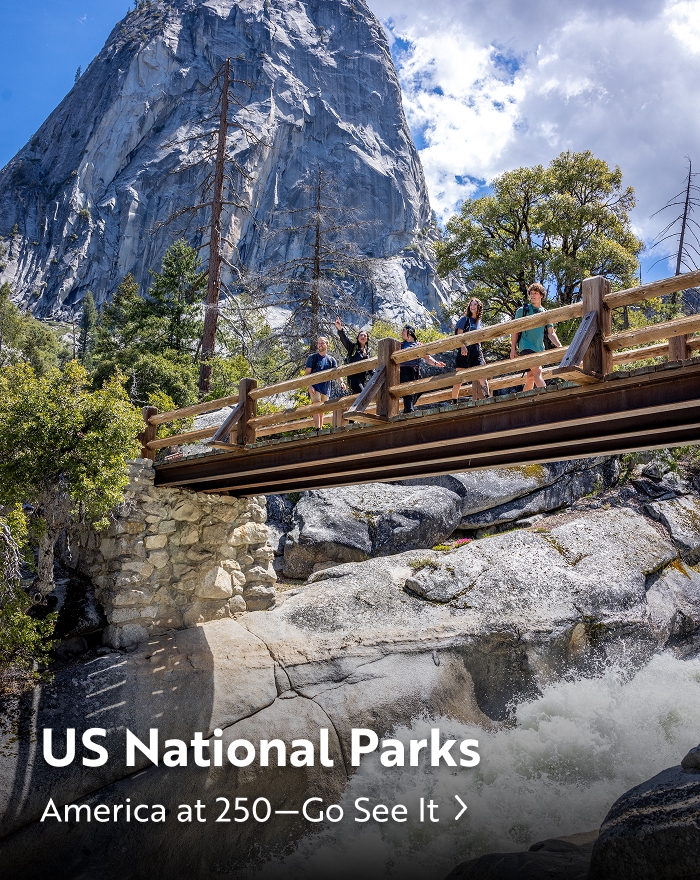 A group of people standing on a wooden bridge over a waterfall with a large rock mountain in the background at a US National Park.