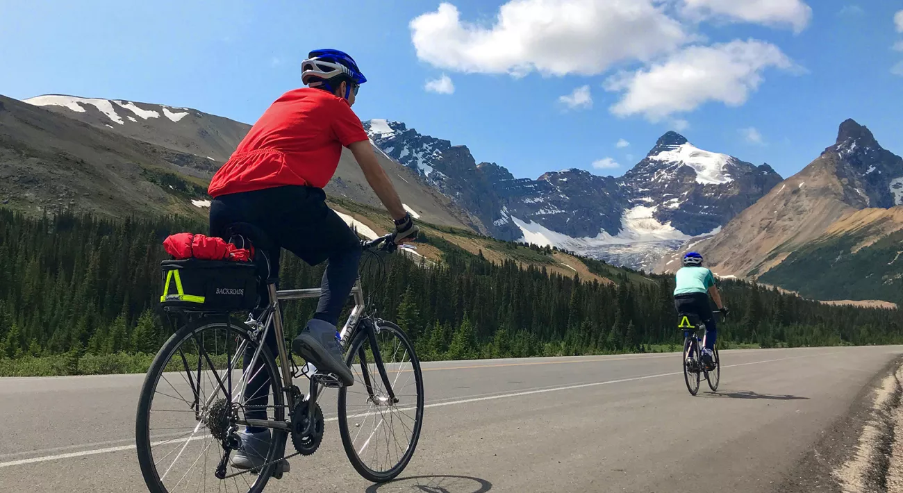 Two guests cycling down mountain road, snowy caps in background.