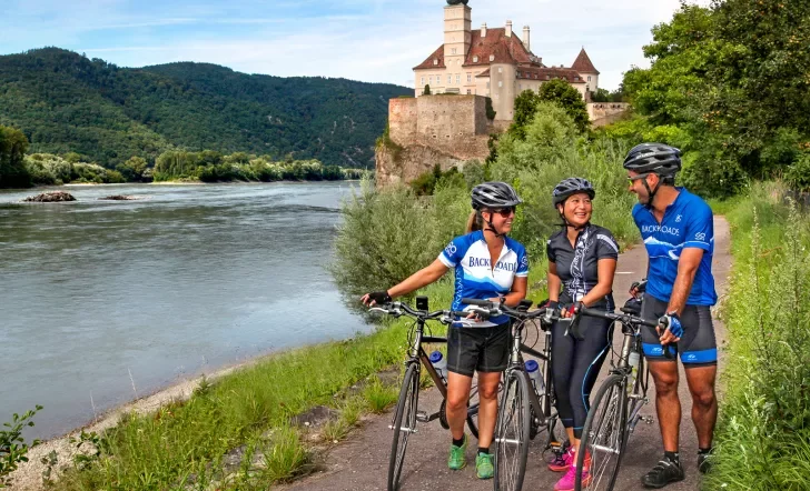 Three bikers walking their bikes on a path along the Danube River.