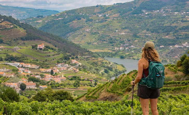 Woman wearing a backpack, standing on top of a hill looking down at a small town surrounded by trees