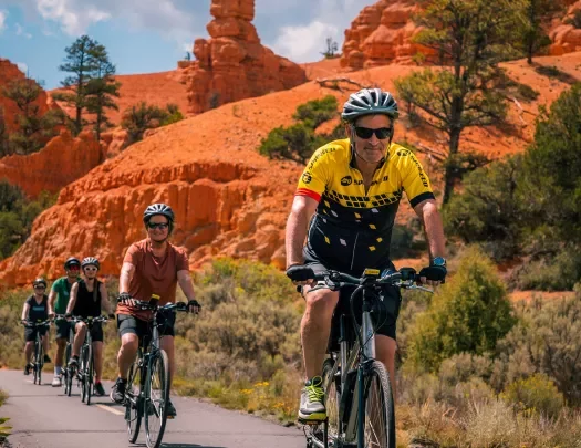 Five guests cycling towards camera, trees, vibrant red sand, hoodoos behind them.