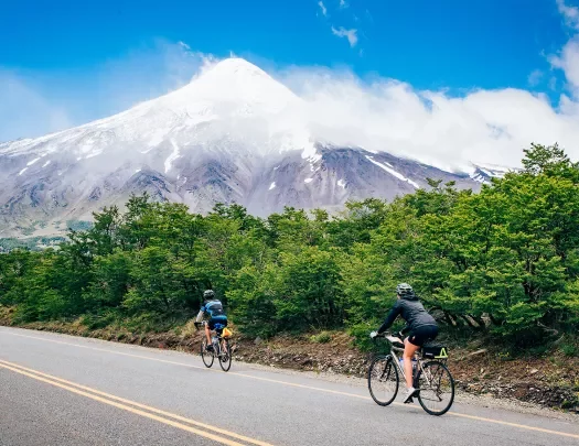 Two guests cycling up road, forest, snowy mountain in distance.