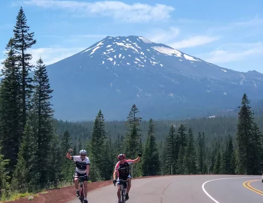 Two guests cycling and waving to camera, Mount Bachelor in background.