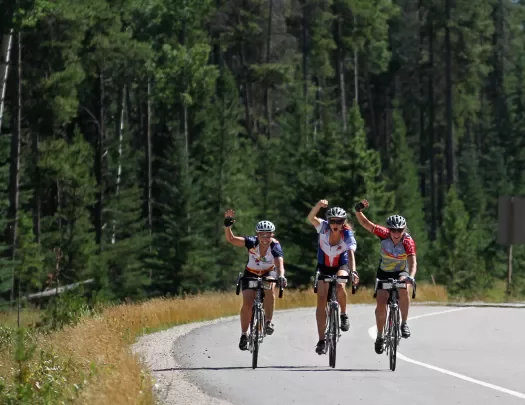 Three guests cycling down large road, waving at camera.