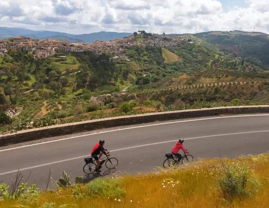 Two guests cycling down road, Medit. hilltop town in distance.