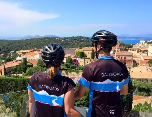 Two guests in bike gear overlooking countryside, tan stone houses, trees all around.
