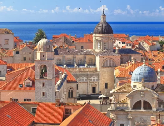 Wide shot of Dubrovnik coastline, blue ocean, white and tan houses.