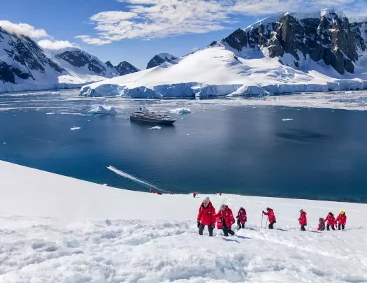 Hikers trekking up a snowy hill in Antarctica with a cruise ship anchored in the background
