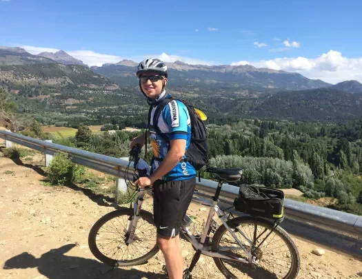 Guest with bike on dirt cliff, overlooking forest, mountains, sky.