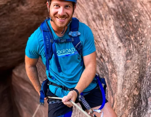 Guest in rock climbing gear on cliffside, smiling at camera.