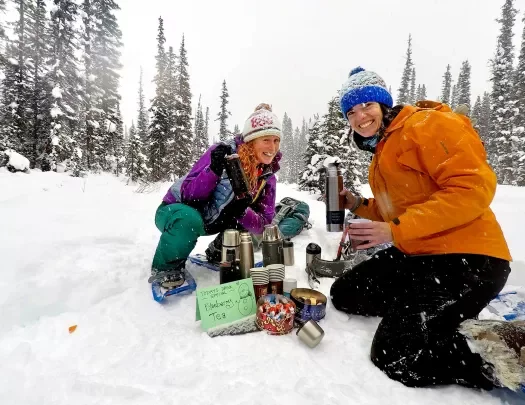 Two guests in snow, getting blueberry tea from thermos.