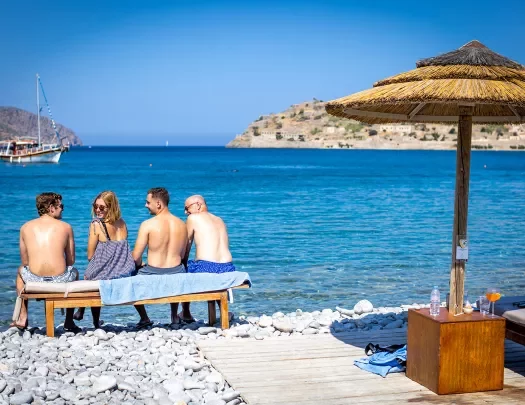 Four guests on rocky beach sitting on bench, overlooking ocean, one looking back at camera.