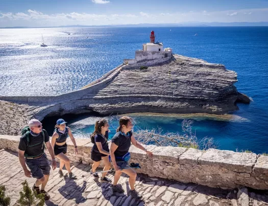 Four guests hiking up rocky staircase, small white lighthouse & ocean in background. 