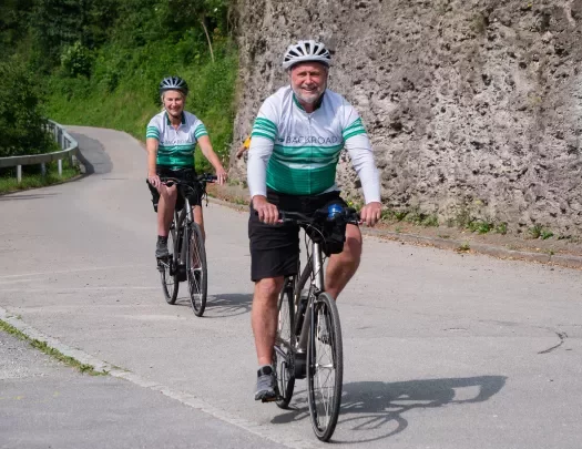 Two guests biking down road, Backroads jerseys on, smiling.