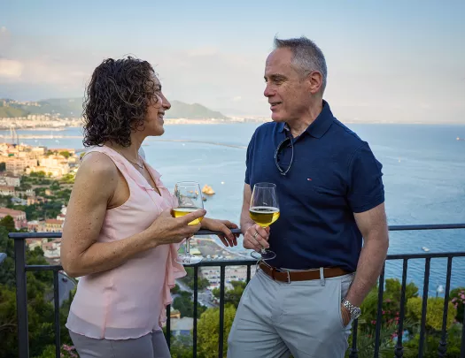 Two guests talking, wine glasses in hand, overlooking coastline.