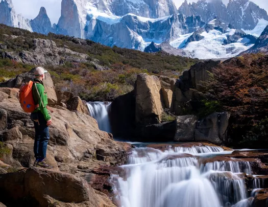 Guest standing next to flowing waterfall, sharp, snowy peaks in background.