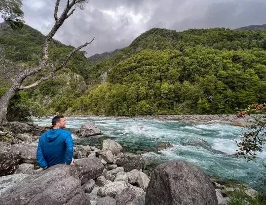 Guest sitting on riverside, white water flowing, mountains and grey clouds above.