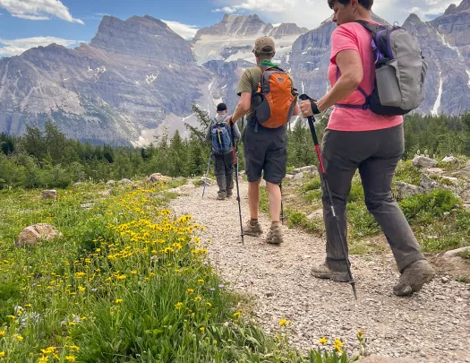 Three guests hiking down meadow trail, Rockies in background.