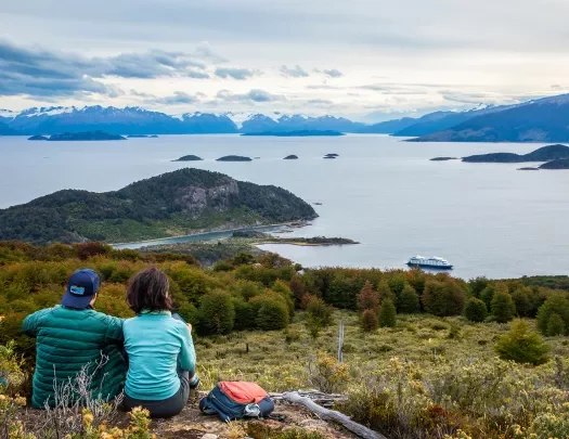 Two guests sitting on hilltop, overlooking huge lake, ring of mountains, clouds, etc.