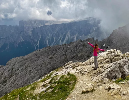 Guest with hiking poles on hilltop, gesturing to large range behind her.