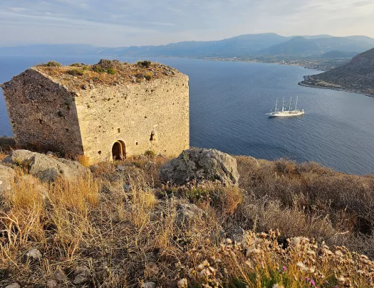 Shot of small stone shack on cliffside, ocean, mountainous coast, boat behind it.