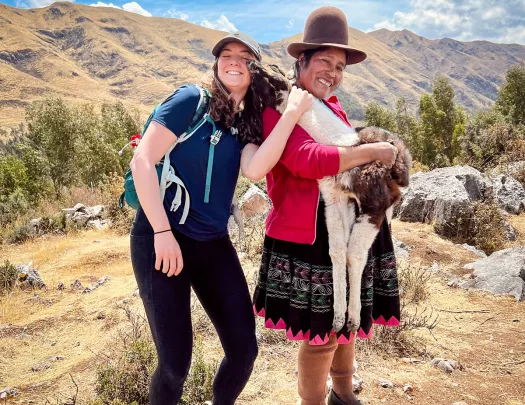 Guest, local woman and baby llama standing together. Llama sniffing guest.