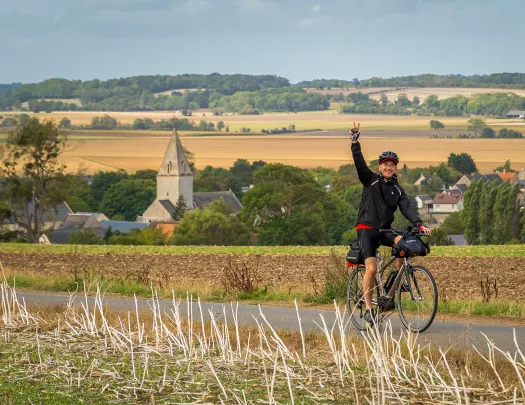 Man biking on an asphalt road in a grass valley, while he's waving his hand in the air