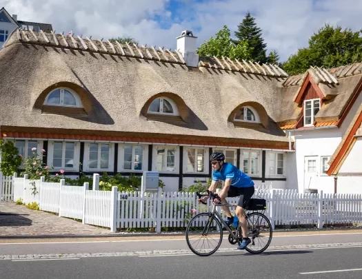 Man biking with a house and a white picket fence behind him