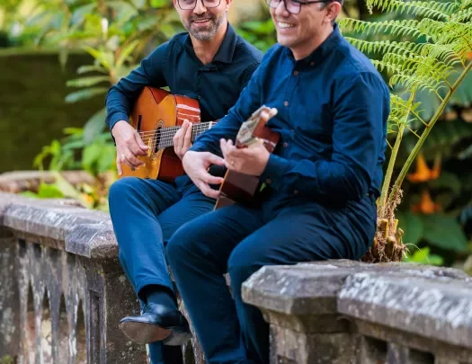 Two men sitting on a stone fence playing guitars and smiling