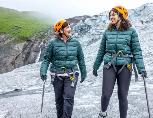 Two women wearing green jackets and orange helmets ascending a snowy hill