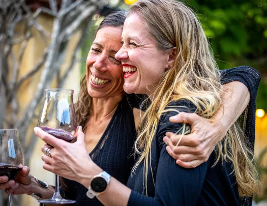 Two women smiling and laughing with arms around their shoulders, holding wine glasses