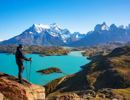 Person with walking poles on top of a rock looking out to a lake and snow-capped mountains