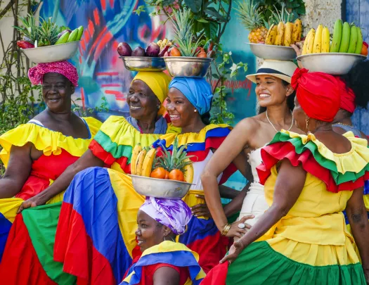 Women in colorful dresses pose with fruit bowls balanced on their heads