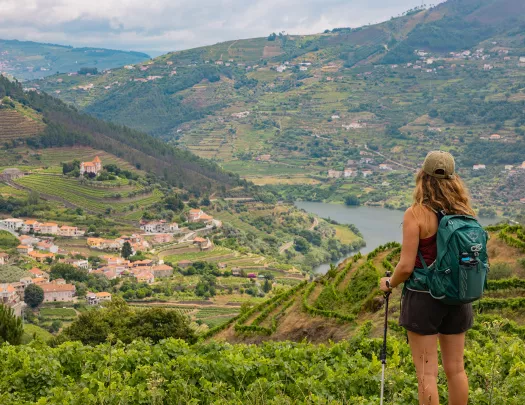 Woman wearing a backpack, standing on top of a hill looking down at a small town surrounded by trees