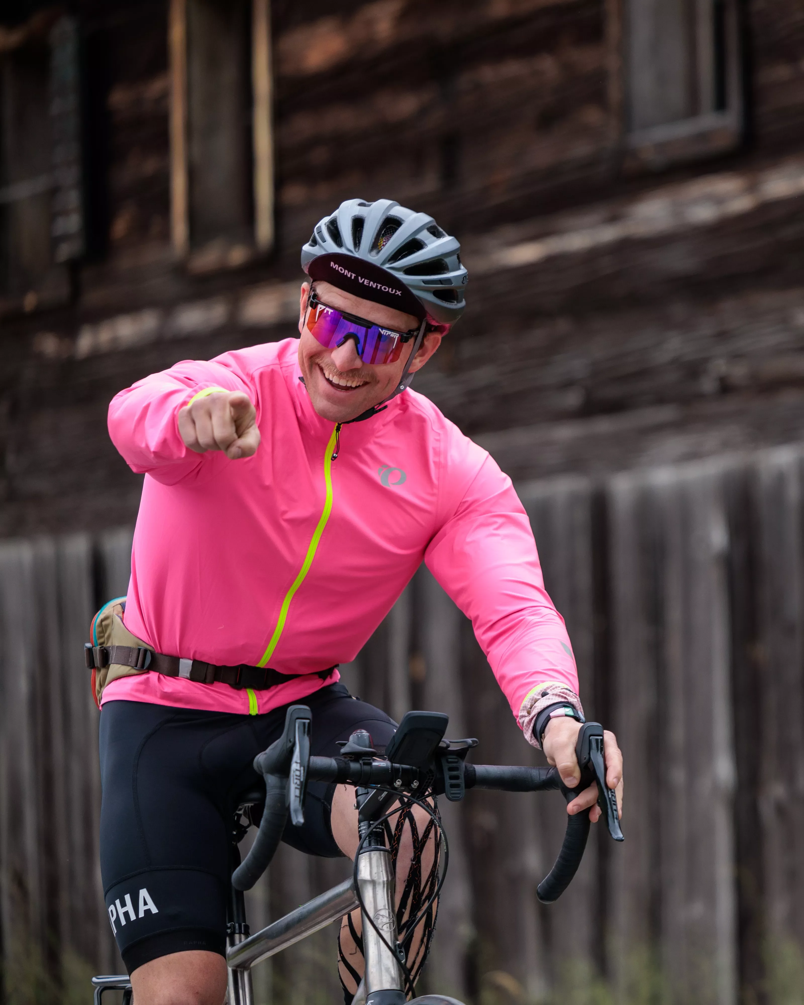 Man with a pink jacket riding a bicycle and pointing at the camera