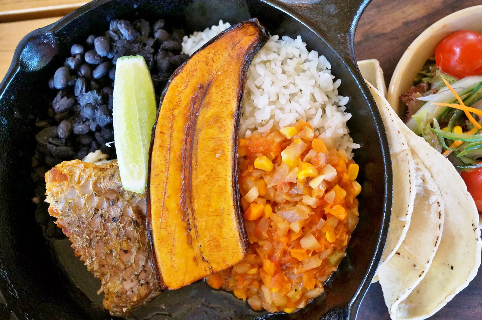 Plate of food with beans, rice, vegetables, grilled fish, and a plantain