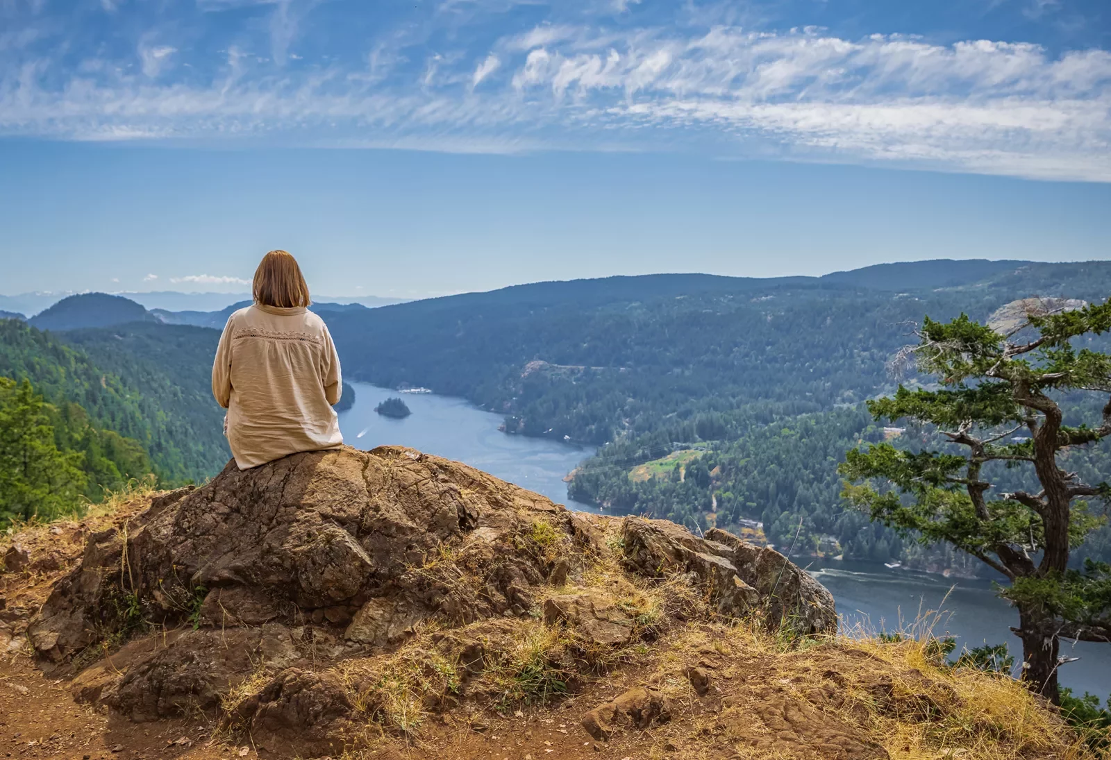 Woman sitting on a large boulder on top of a hill, looking down at the water