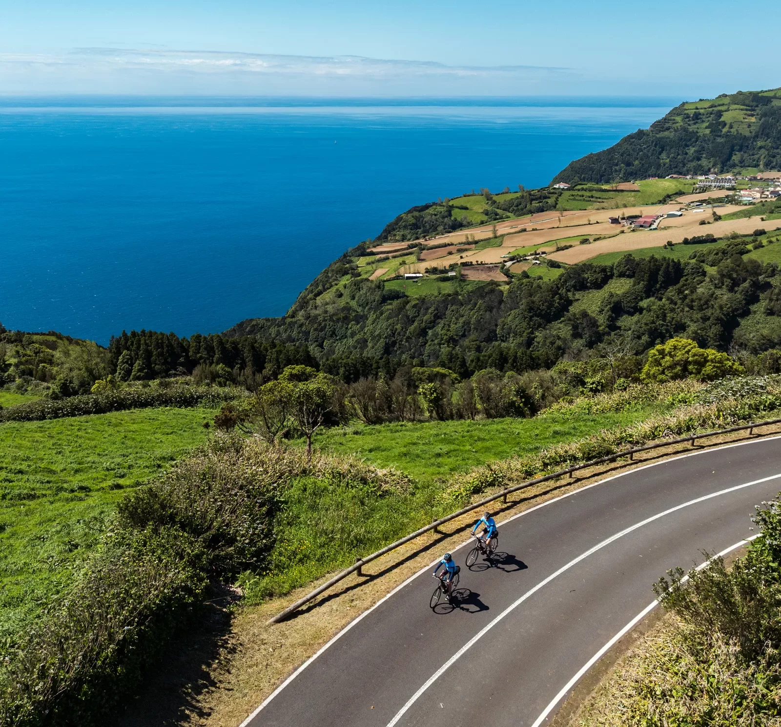 Two people riding their bikes on an asphalt road with a large valley and ocean in the background