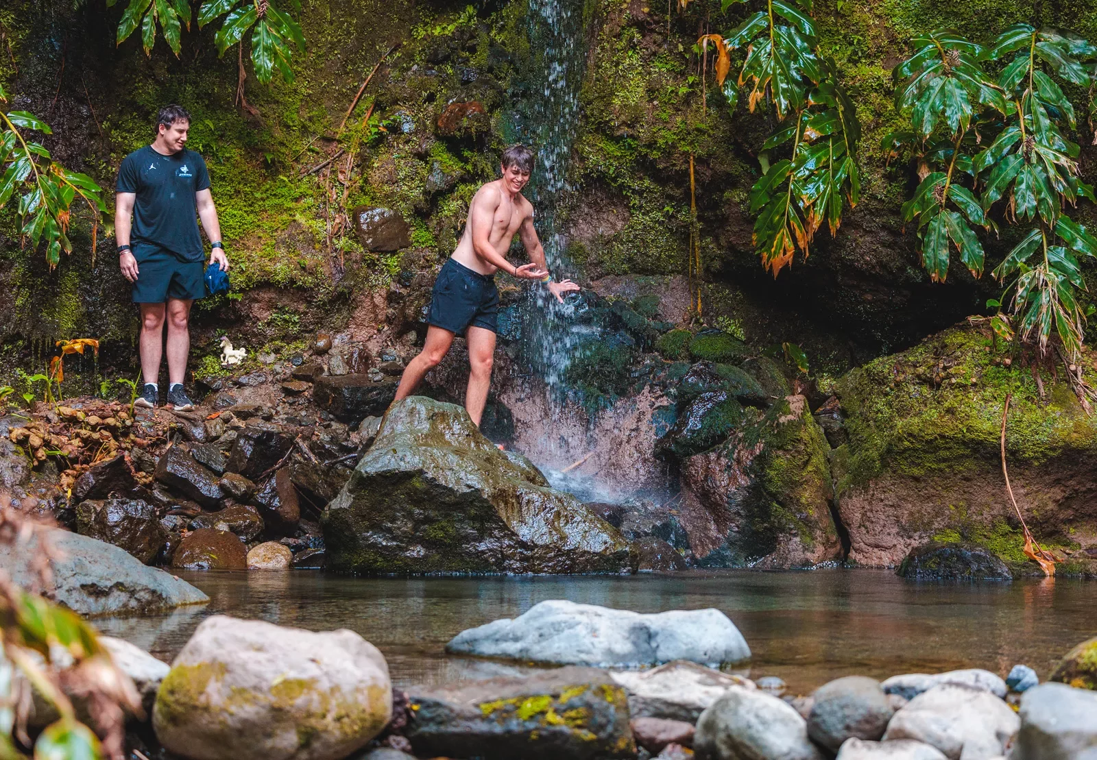 Two men standing on wet rocks under a waterfall, preparing to jump into a lake