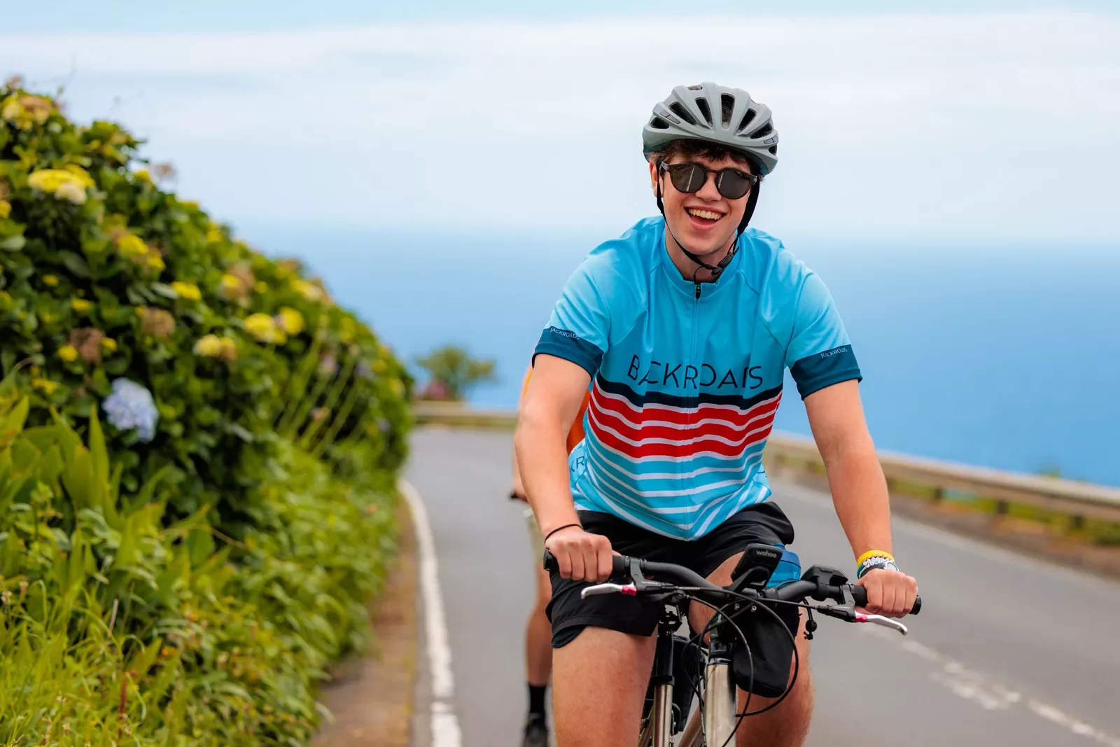 Man with a blue jersey and sunglasses riding a bike on a road next to the ocean