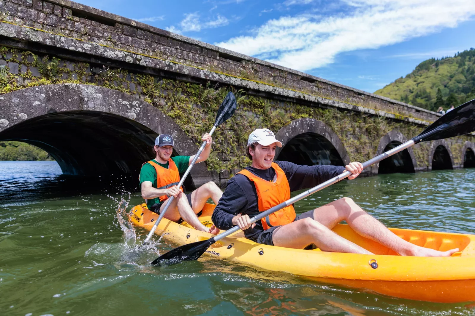 Two men on a kayak, paddling in a river by a bridge
