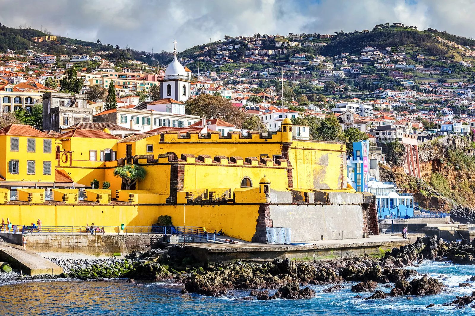 Yellow building by the coast of an ocean, with a hill of houses in the dstance