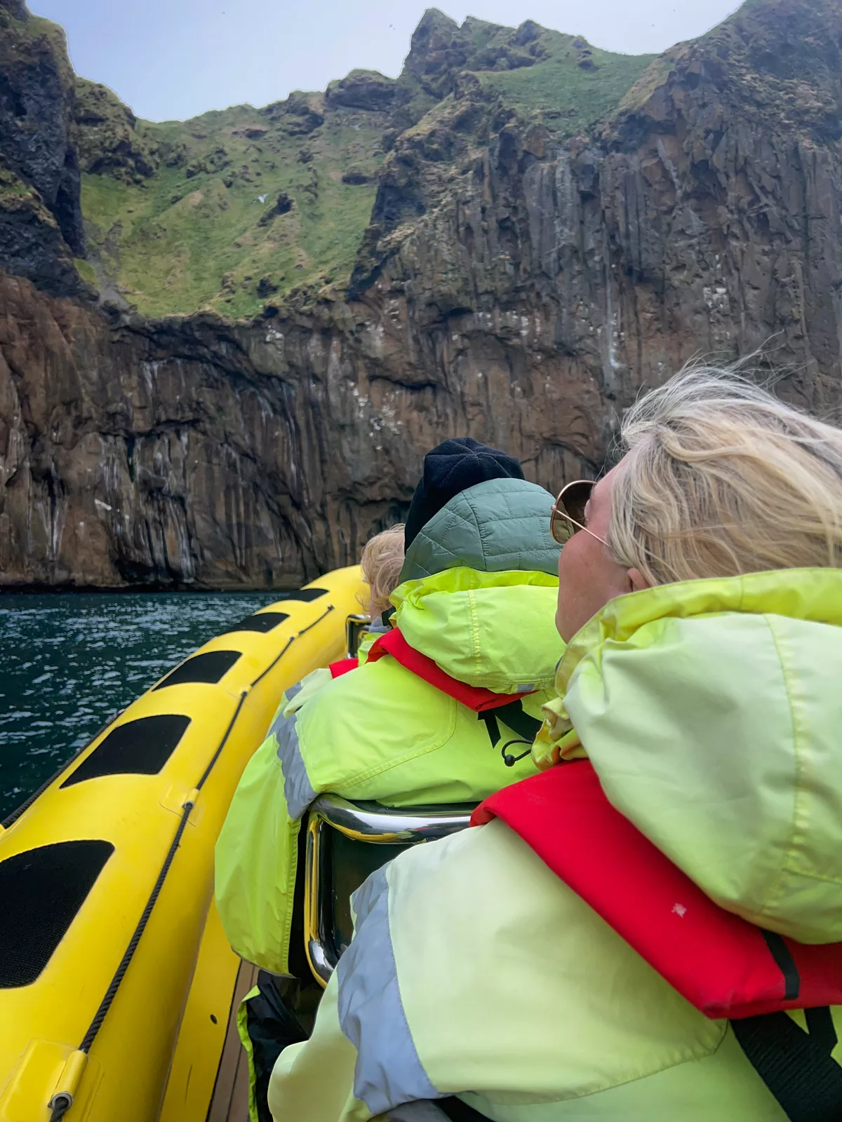 Group of people on a yellow raft, wearing green jackets going towards a cliff