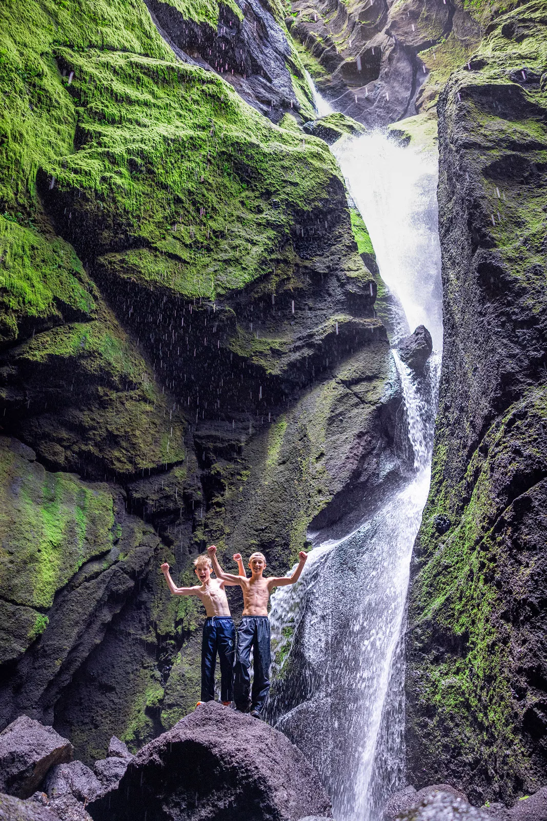 Two boys standing on a rock next to a waterfall