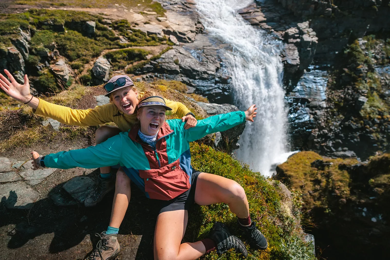 Two women with their arms open on a cliff with a waterfall below