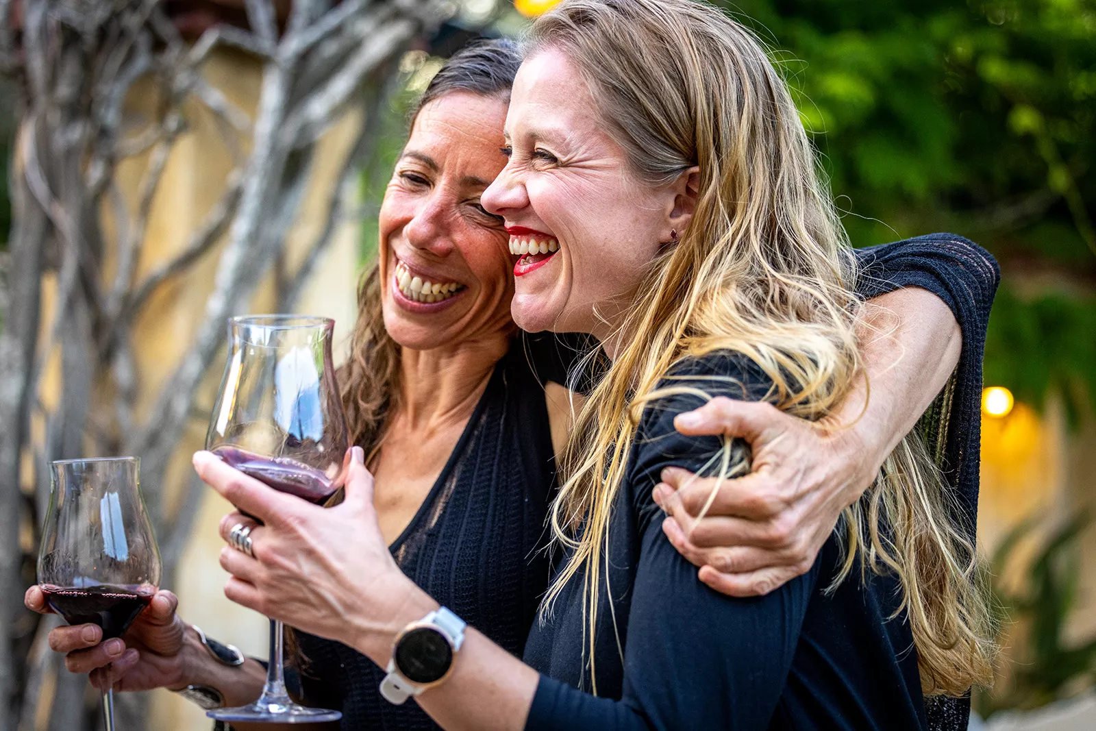 Two women smiling and laughing with arms around their shoulders, holding wine glasses