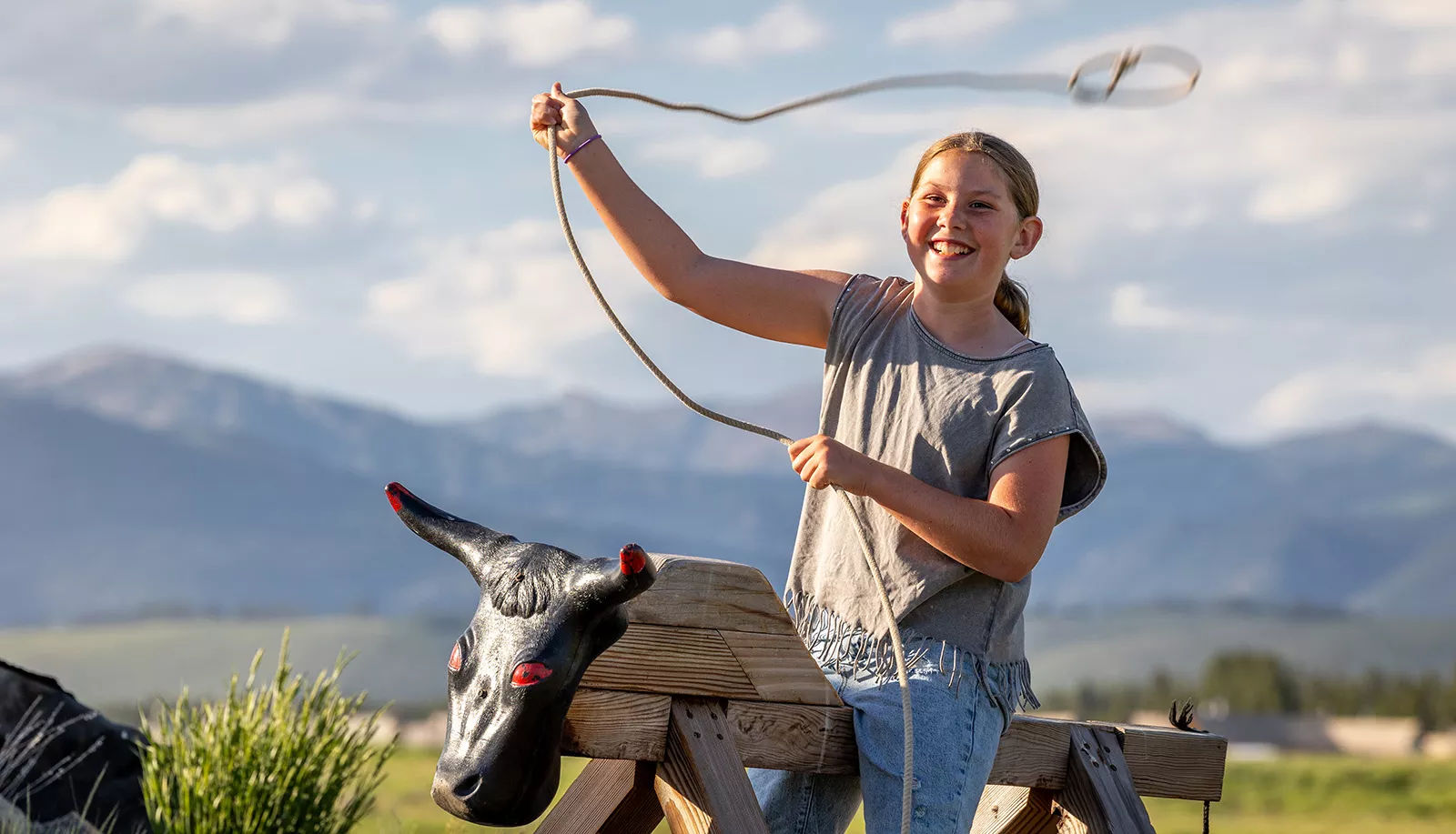 Girl riding a wooden bull prop while swinging a rope