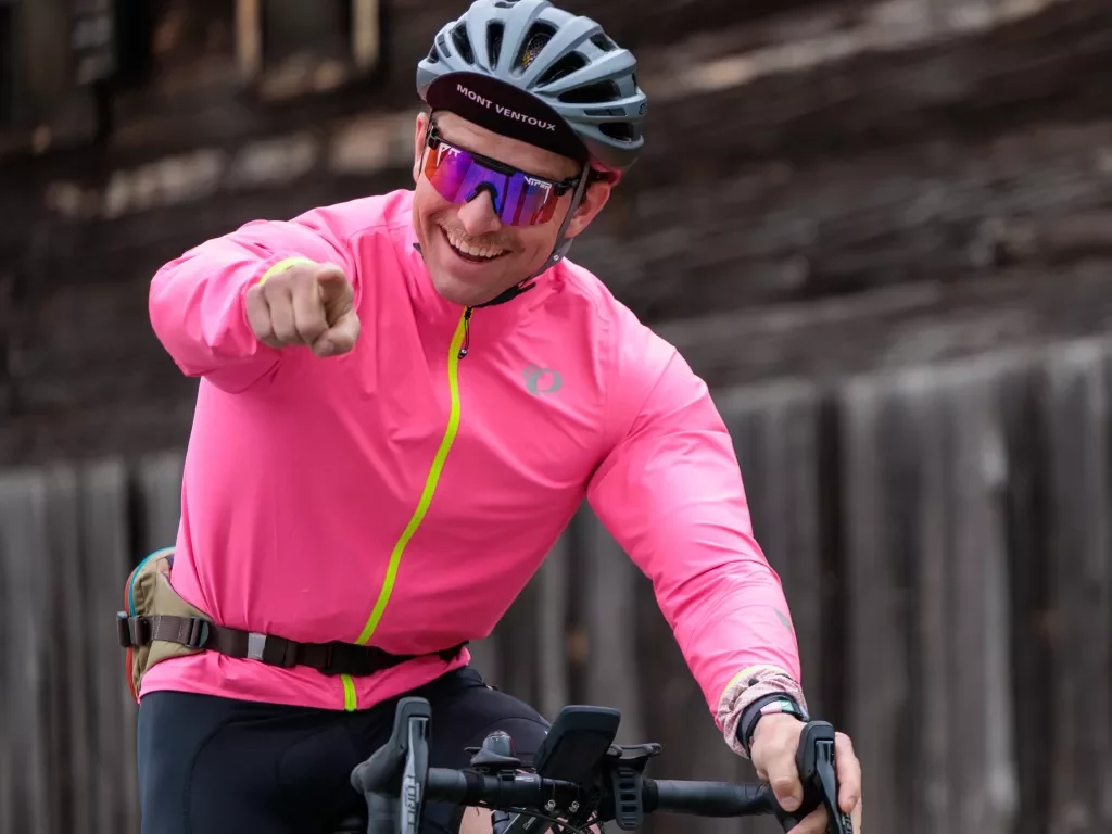 Man with a pink jacket riding a bicycle and pointing at the camera
