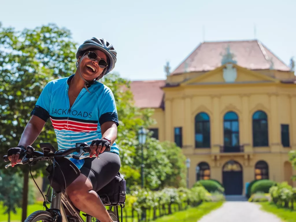 Woman wearing sunglasses and riding a bike in front of a long pathway leading to a yellow building