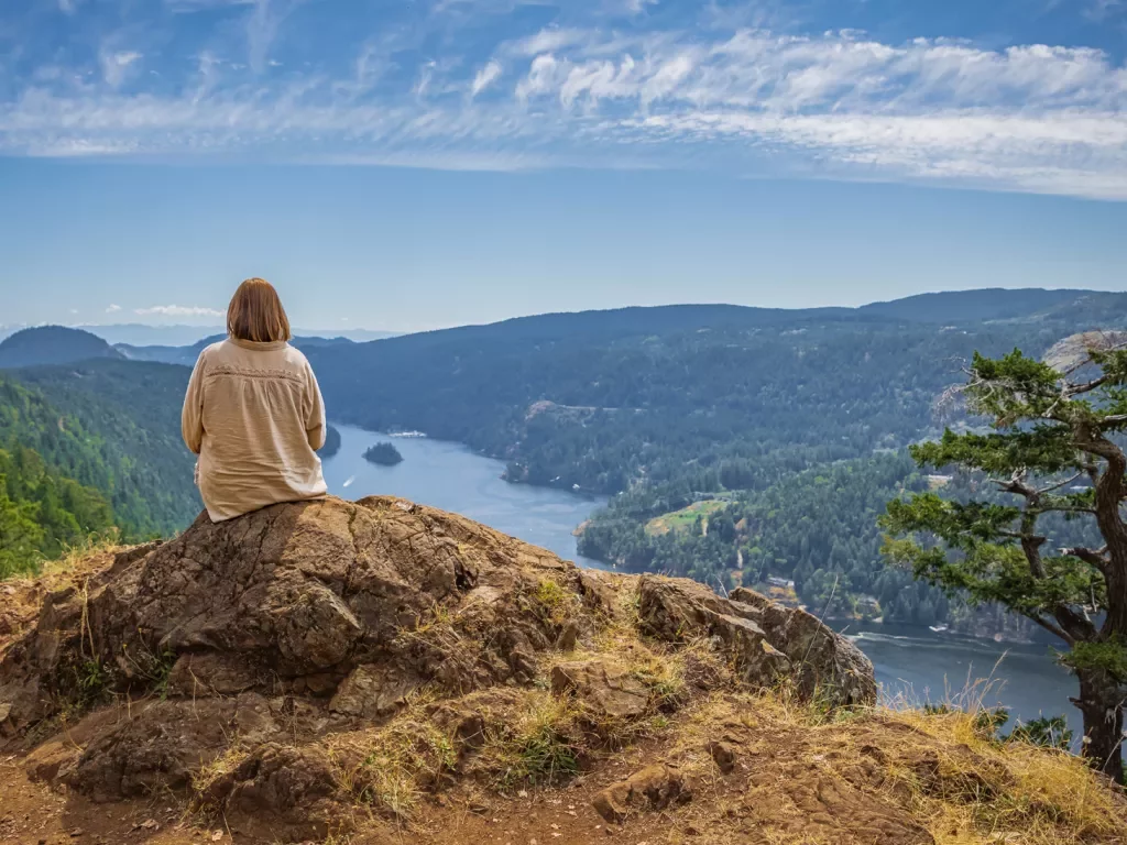 Woman sitting on a large boulder on top of a hill, looking down at the water
